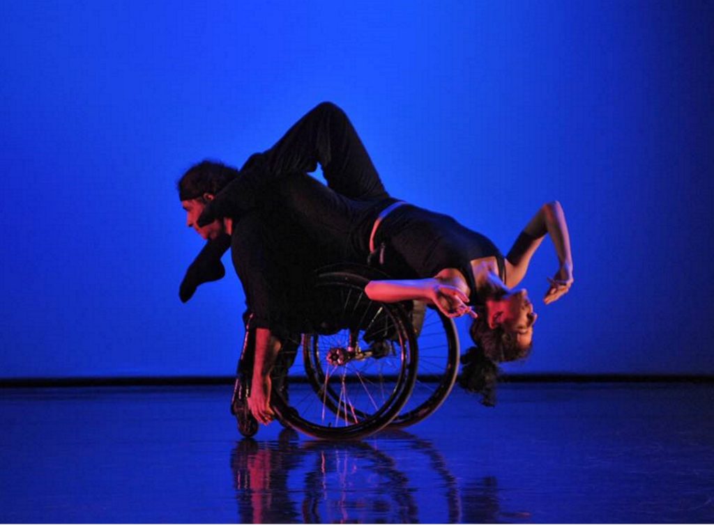 White female dancer balances upside down on the back of a white male dancer who uses a wheelchair