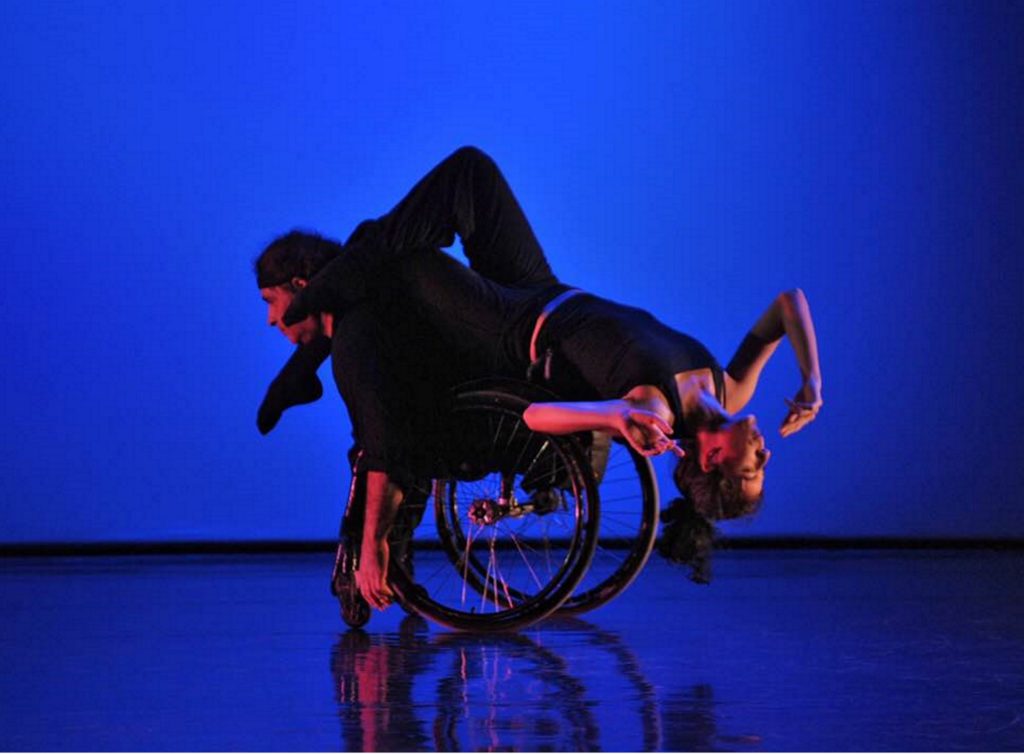 White female dancer balances upside down on the back of a white male dancer who uses a wheelchair