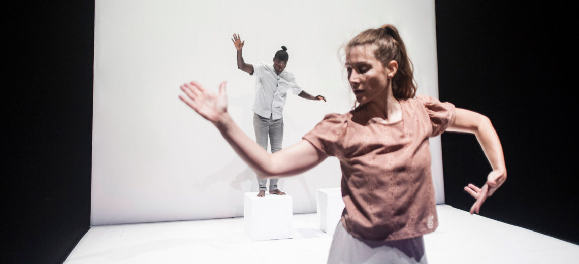 On a white spotless stage, two dancers open their hands like if trying to find their balance, one in the very front of the stage and one in the back on a white cube.