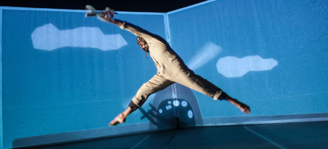 A dancer in a pilot's suit holding a toy airplane in his hand, jumps up hi, in front of a background representing an open book with painted clouds and a cockpit.