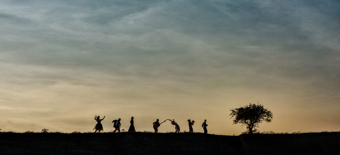 7 dark strangely shaped human silhouettes walk on the top of a dark hill towards a tree in front of a cloudy colourful sky. 