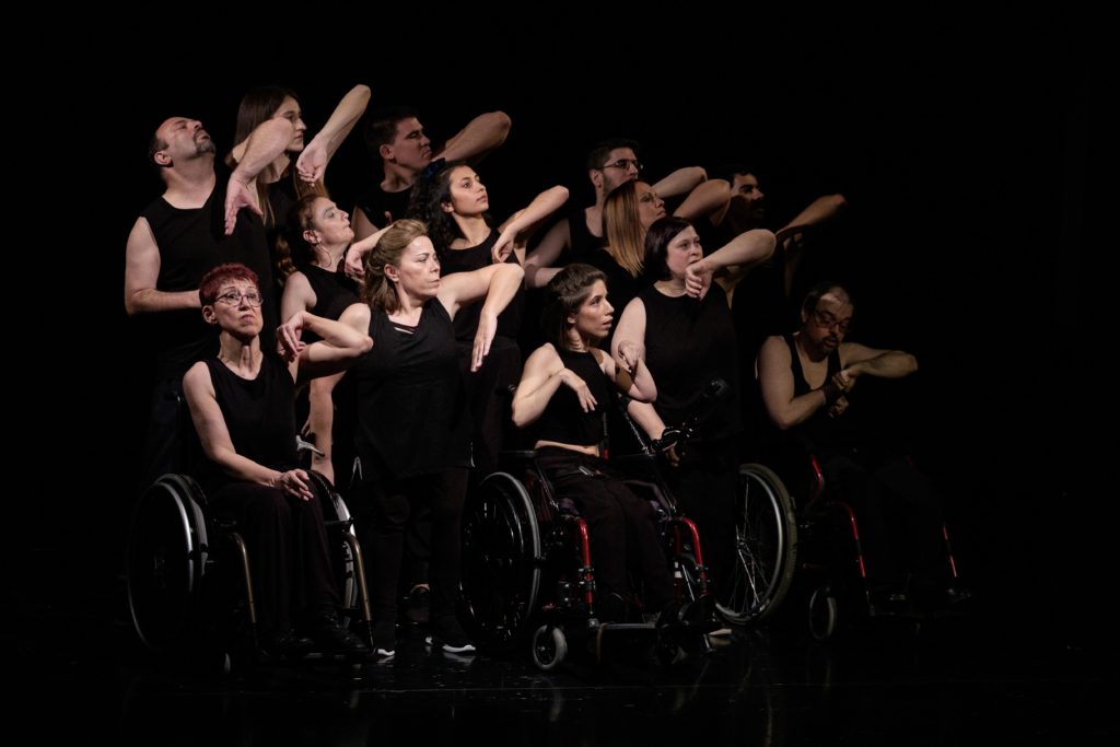 Ensemble of white disabled and non-disabled dancers all dressed in black, they are raising their left elbows in unison.