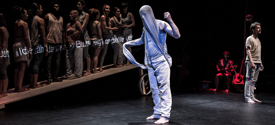 A performer with his head covered by an elastic blouse wrapped around him, stands in the middle of the stage while behind him, 10 other performers stand next to each other on a sloping wheeled table.