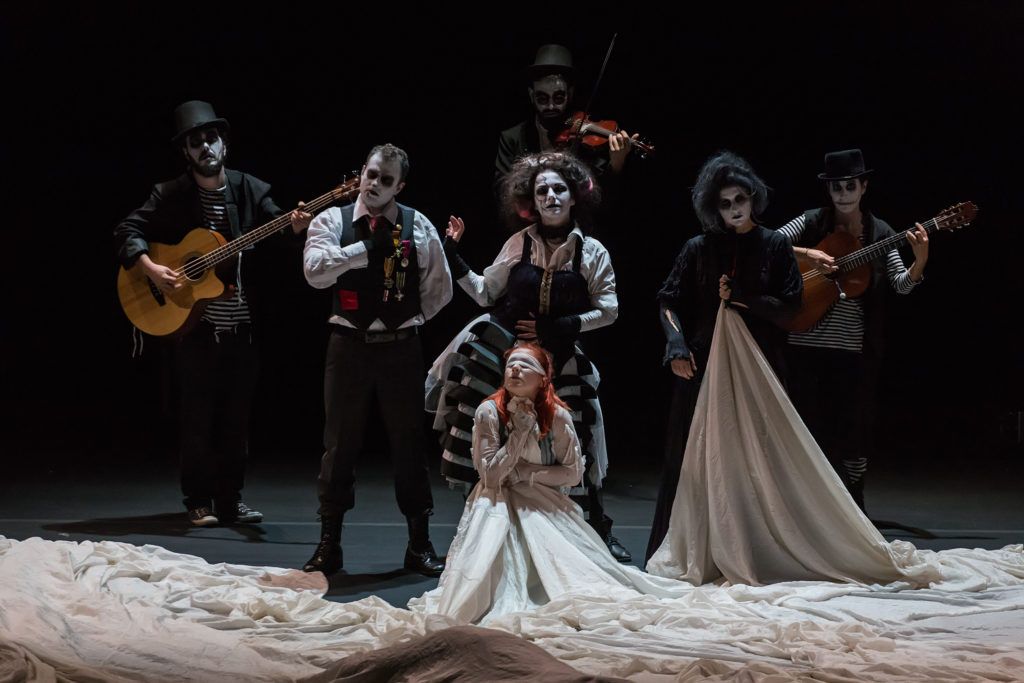 6 performers in black and white face paint play instruments whilst another in the centre of them is blindfolded, wearing a dress with a massive train which is bunched up around them all.