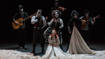 On a blackbox stage, 6 performers with white painted faces and black eyes, dressed in old-used aristocratic costumes and caring instruments are looking towards us, whilst another in the centre of them is blindfolded, wearing a white massive dress with its tail bunched up around them all.
