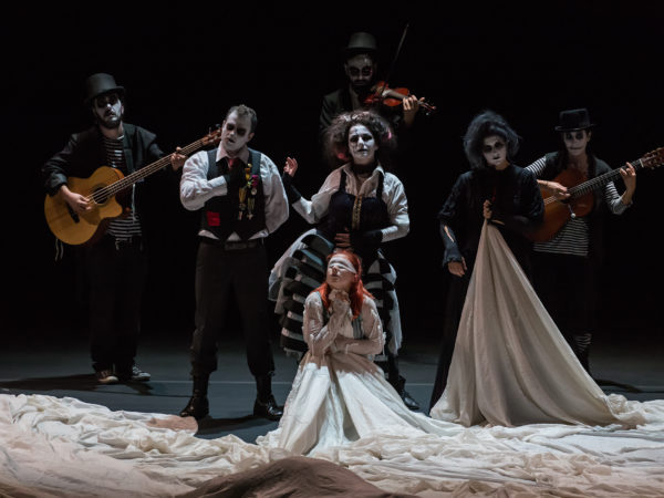 On a blackbox stage, 6 performers with white painted faces and black eyes, dressed in old-used aristocratic costumes and caring instruments are looking towards us, whilst another in the centre of them is blindfolded, wearing a white massive dress with its tail bunched up around them all.