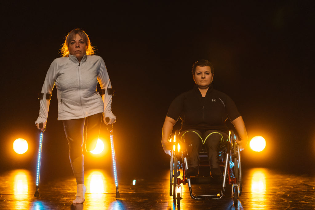 Two white female dancers on stage. One is in a wheelchair and the other is using illuminated crutches. They are brightly backlit.