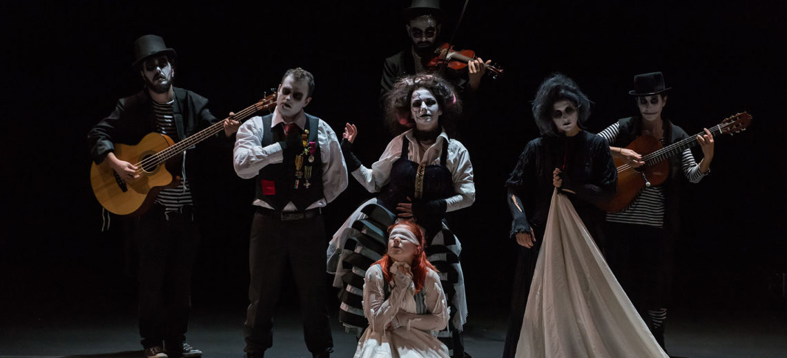 On a blackbox stage, 6 performers with white painted faces and black eyes, dressed in old-used aristocratic costumes and caring instruments are looking towards us, whilst another in the centre of them is blindfolded.