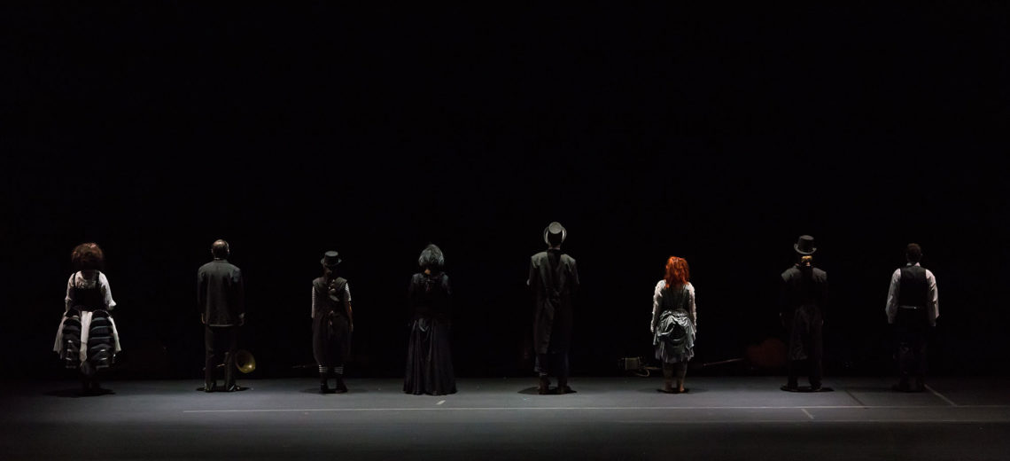 8 performers stand in line at the end of a black stage with their back towards us, dressed in black old fashion used aristocratic costumes and top hats.