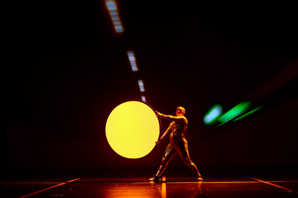 A learning disabled man ina  silver body suit holds a glowing yellow circle on a stage