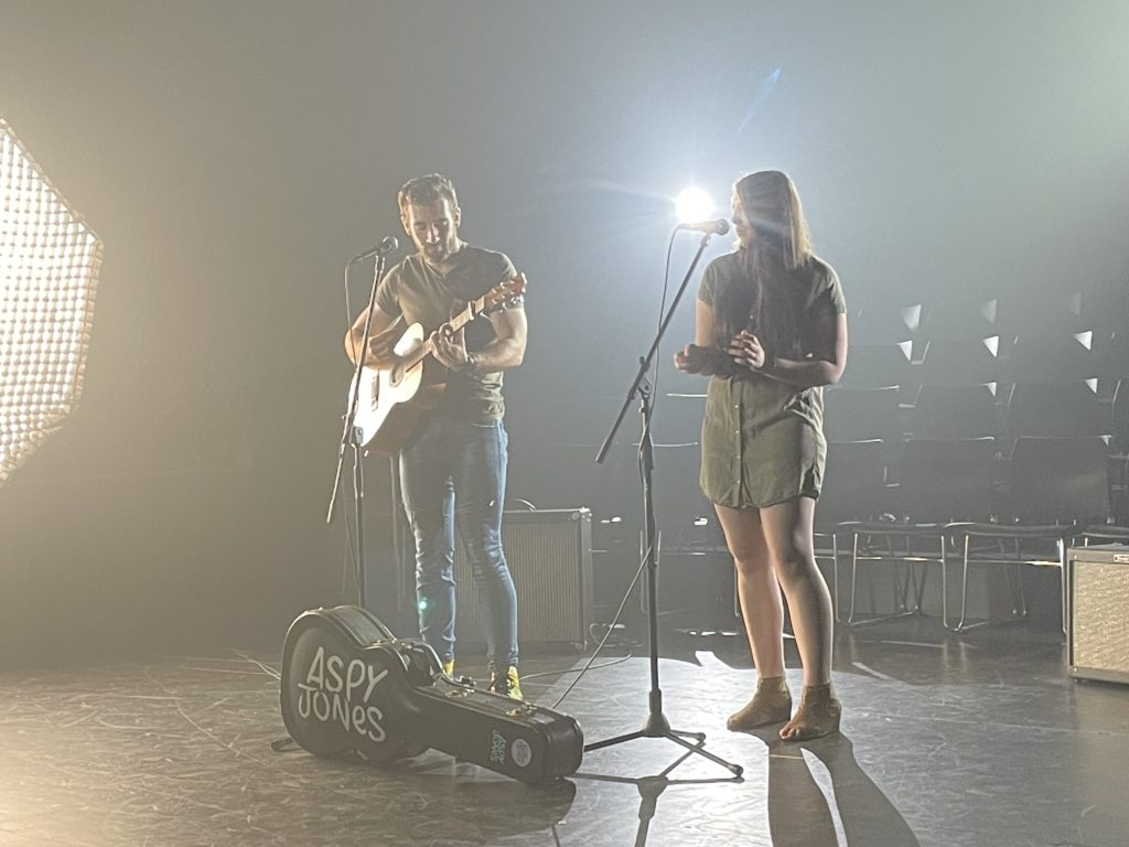 A white female singer with a white male guitarist stand on stage performing