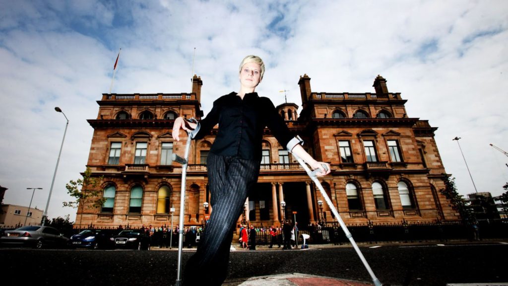 A white woman with crutches stands in front of a grand Victorian building