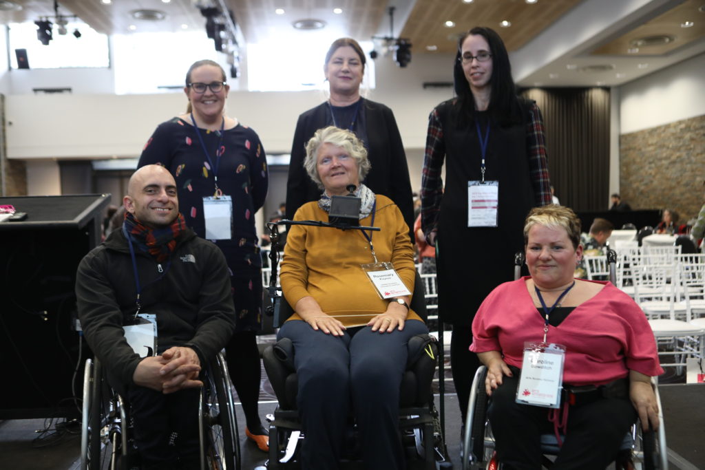 6 disabled people, three in wheelchairs smile for the camera, each with large name tags around their necks