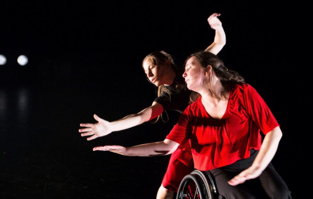 A female dancer in a wheelchair wears a bright red top contrasting on a black background.