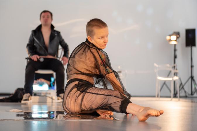 A woman with a shaved head, clothed in a meshed semi-transparent outfit stretches one leg across the floor.