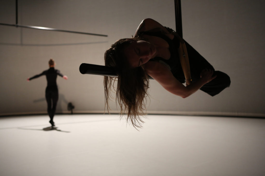 A white female dancer with a prosthetic arm balances on a floating L-shaped pole, while another dancer dressed in black performs in the background.