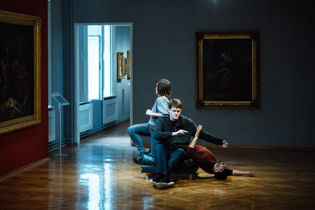 Three performers pile together in an art gallery, contorting their hands and grimacing