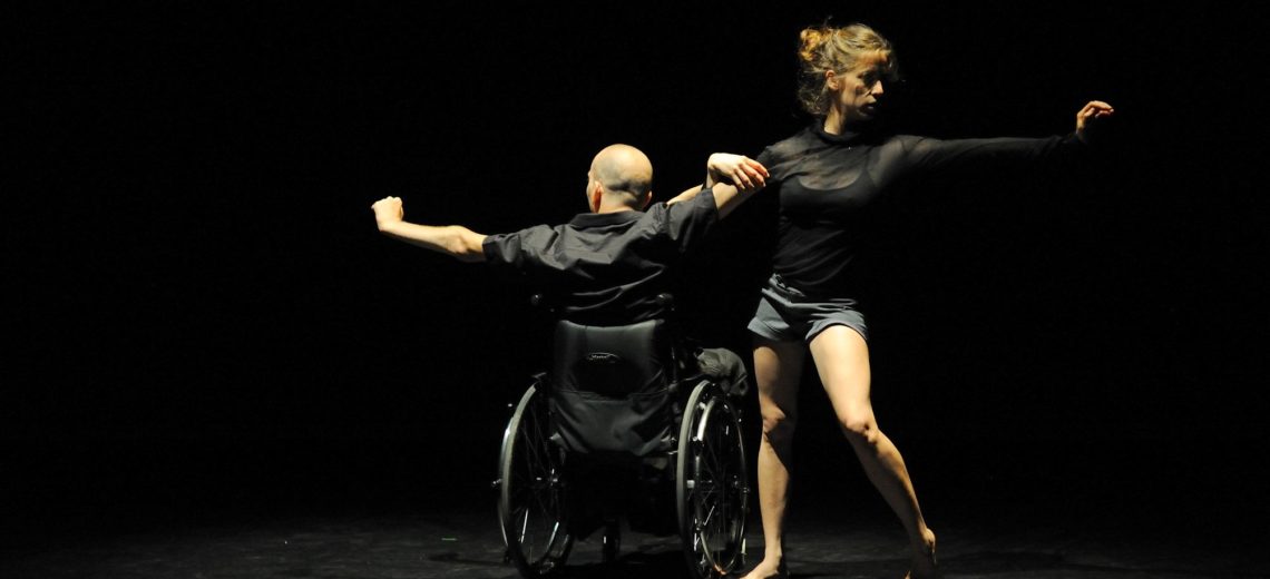 Woman dancer next to Male wheelchair dancer, both are reaching out with one hand while the other hands are intertwined 
