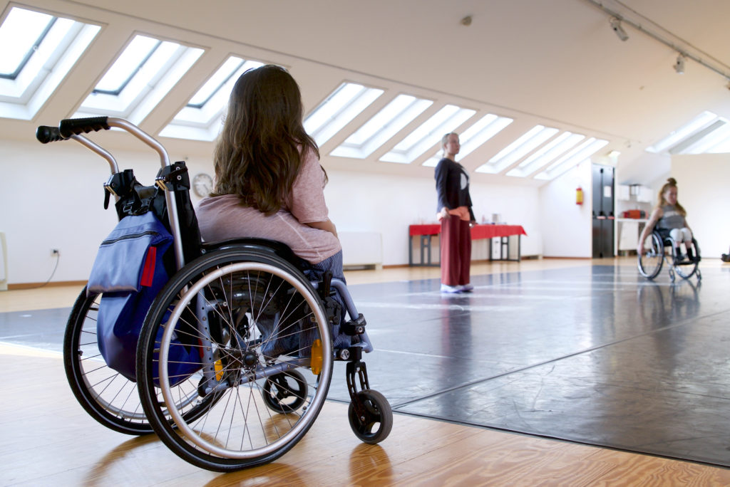A female choreographer in a wheelchair sits directing a group of dancers in a bright studio