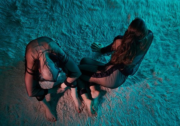 Two bodies from above bathed in bluish-bronze light, on a soft flokati carpet. They are wearing transparent tracksuits, their skin color is white, they are barefoot. The person on the left rests their arms on their drawn knees, holds their left wrist with their right hand. The head falls between the legs. The person next to them lies on their right side. With the bent legs crossed and leaning on their hands, they look forward to the right - beyond the picture. They touch the other with the tips of their feet. One remains, the other seems to leave the place in the next moment. Long dark hair, no hair, black mesh on skin.