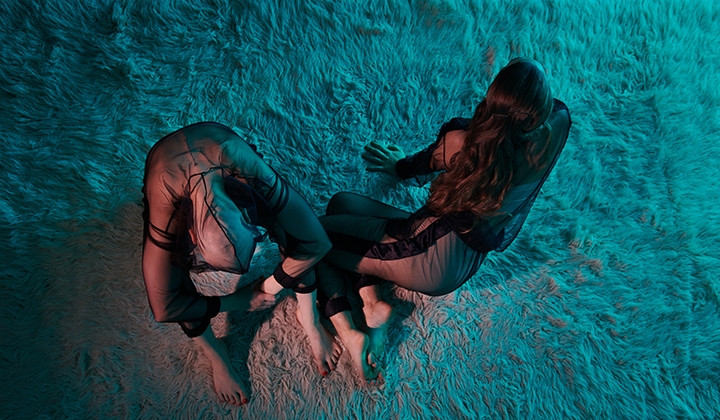 Two bodies from above bathed in bluish-bronze light, on a soft flokati carpet. They are wearing transparent tracksuits, their skin color is white, they are barefoot. The person on the left rests their arms on their drawn knees, holds their left wrist with their right hand. The head falls between the legs. The person next to them lies on their right side. With the bent legs crossed and leaning on their hands, they look forward to the right - beyond the picture. They touch the other with the tips of their feet. One remains, the other seems to leave the place in the next moment. Long dark hair, no hair, black mesh on skin.