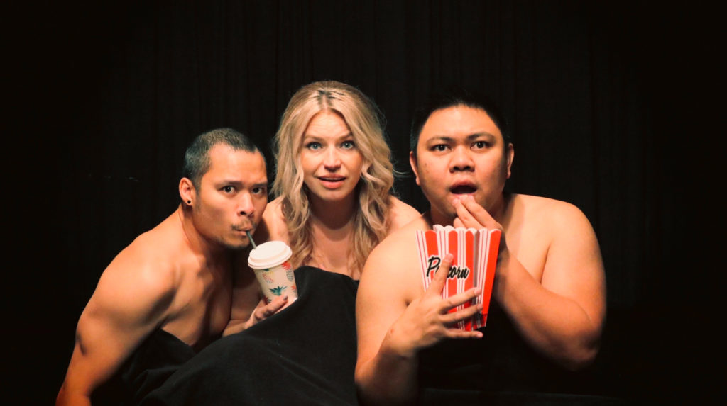 Three artists stare at us like at a film, naked under a black curtain. On the left is a Filipino-Canadian man with black hair and moustache, sipping from a straw in the drink of a blonde woman of Polish background to his right. To her right, a Filipino man holds a cardboard box of popcorn in one hand, his other at his mouth in surprise or fear.