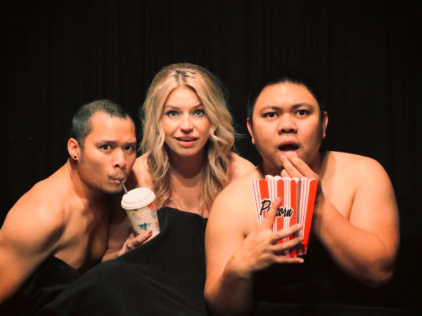 Three artists stare into the camera as if watching a film. They're naked but covered by a black cinema curtain. On the left is a man from Fairford Reserve with short black hair and a moustache. He drinks from a straw from the cinema-style drink held by a blonde white woman of Polish background to his right. On her right, a Filipino-Canadian with short black hair holds a cardboard popcorn box in one hand, while his other hand is at his mouth in surprise or fear.