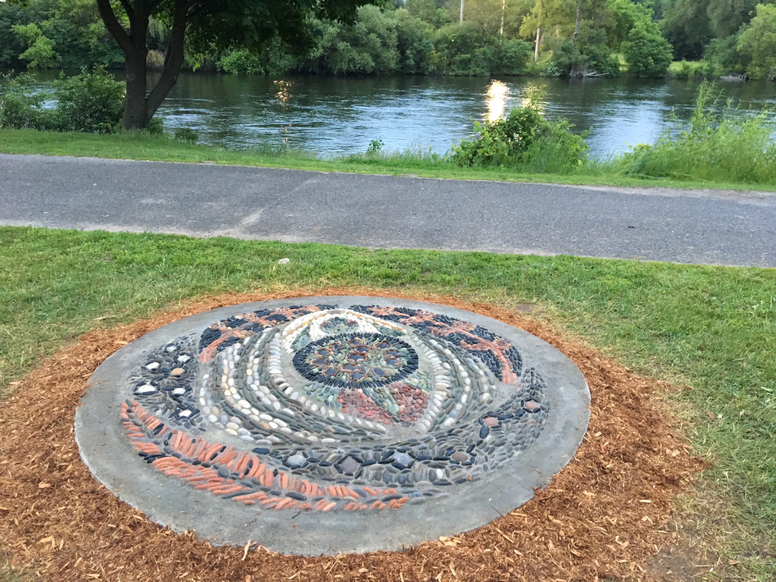 A two metre in diameter stone mosaic is in the foreground. The mosaic features the image of a turtle (Turtle Island), surrounded by cedar trees and water. It's made of red, black, white, tan and grey stones, and surrounded by cedar mulch and grass. In the background, an asphalt path, the Otonobee river, green grass and trees.