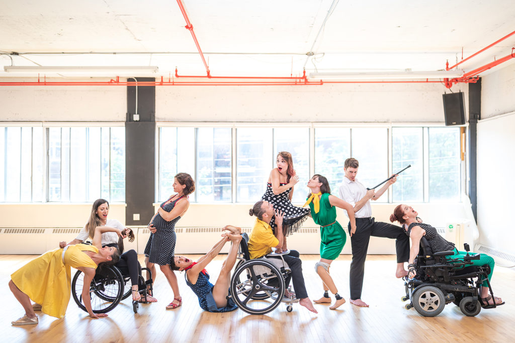 Dance troupe in a dance studio lined up. The dancers are white with a range of imapairments including wheelchair users and amputees. Their is also a pregnant woman holding her bump.