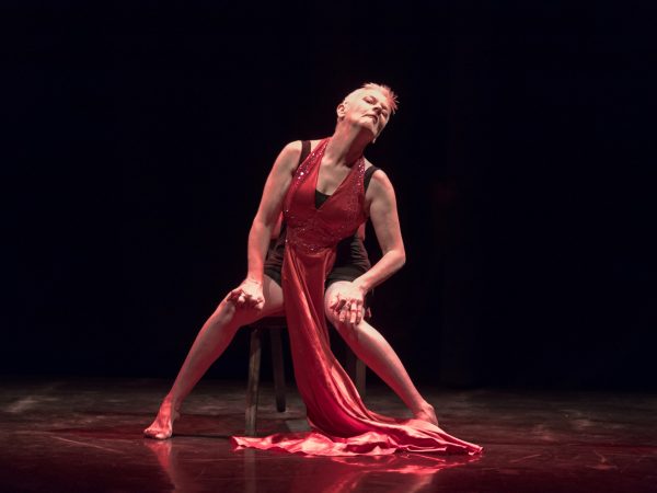 There are two dancers. One is standing in front, wearing a short red dress and she is staring down towards the camera lens. One arm is by her face, and the other by her side. The other dancers is perched on a chair behind, dressed in a light green dress. We see half her face. She is crossing her legs, and in a poised, reclining position. The photo is taken in a bright studio. 