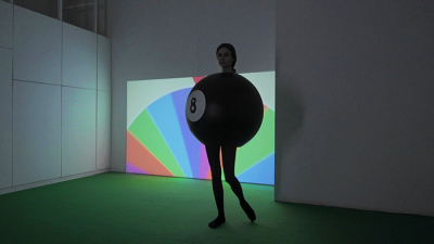 A performer in black dressed dressed as a billiard ball, in front of a colourful spinning wheel