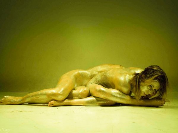 A gold-tinted photo of a female nude performer lying on the ground, covered in glitter