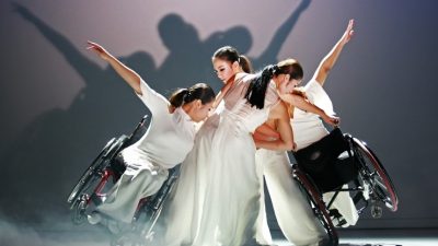 East Asian dancers in white clothing huddle together with two wheelchair users on the edge of the huddle leaning in