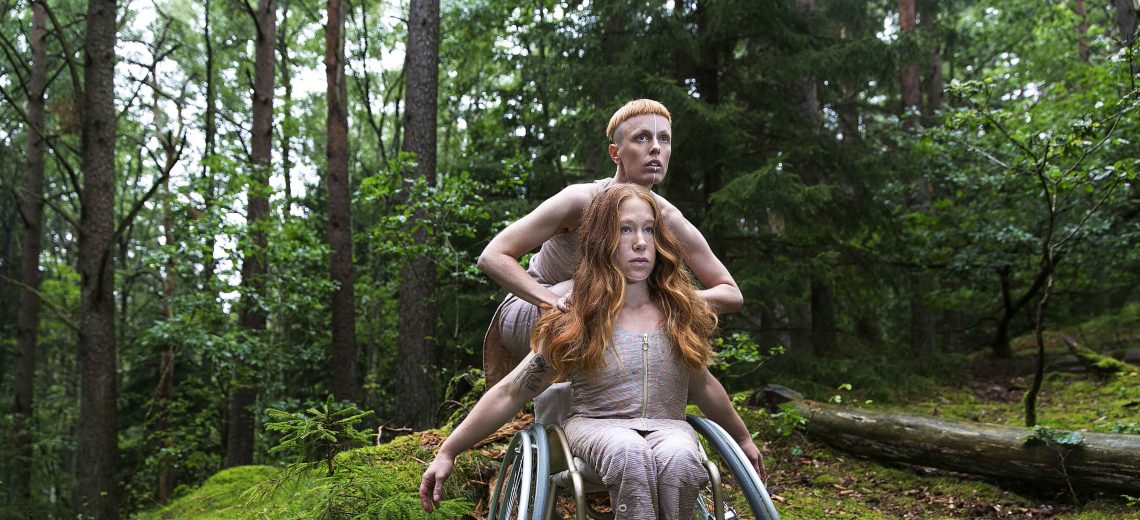 Two dancers are in forest. One dancer is in a wheelchair and the other dancer is squatting behind. 
