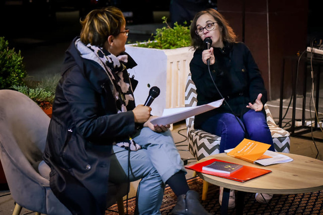 Natalija sits speaking passionately into a microphone, being interviewed by a journalist.