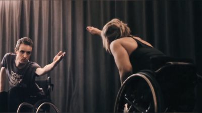 A male and female duo using wheelchairs reach their hands across to join each other