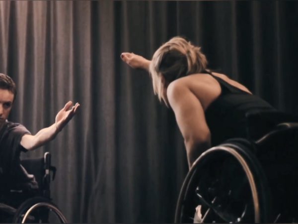 A male and female duo using wheelchairs reach their hands across to join each other