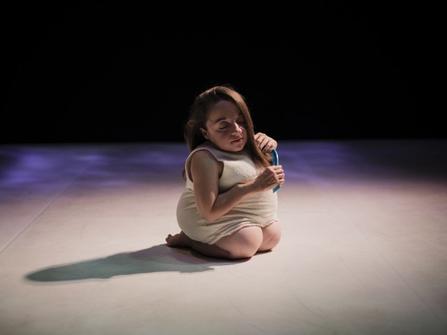 A white woman of short stature kneels on stage stroking her hair