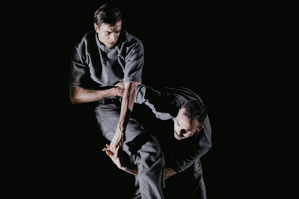 Two white male dancers in grey outfits link arms underneath one of the dancer's legs