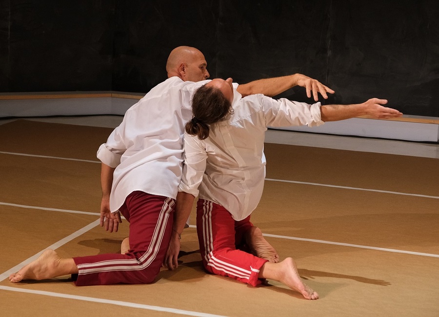 Two male dancers in tracksuits kneel while one brushes over the other