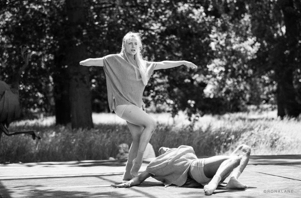 A disabled white female performer with blond hair balances on one leg above a man writhing on the floor. A black and white photo of a performance outside.