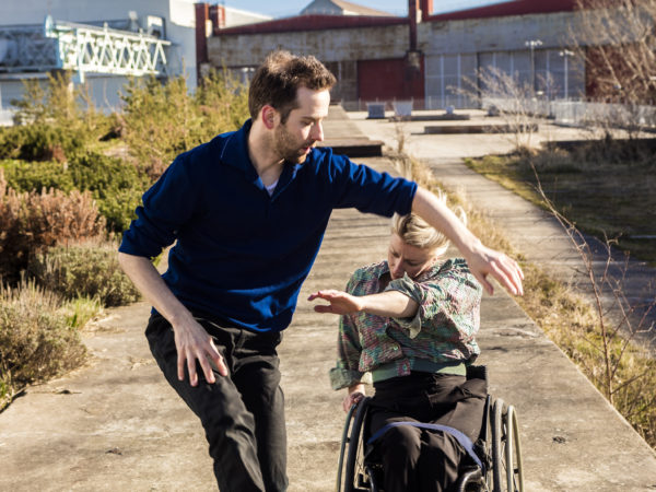 A female dancer with blonde hair using a wheelchair duets with a non-disabled male dancer outside in the sun.