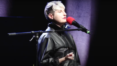 A performer with cropped bleached hair speaks passionately into a microphone