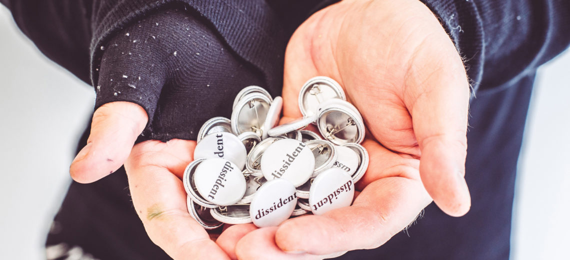 hands holding a number of badges with dissident printed in black ink on a white background