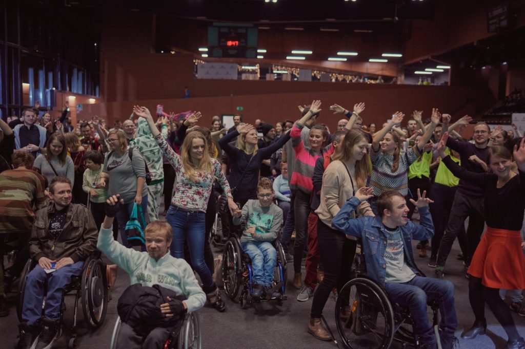 A group of people in a hall, cheering and waving hands above their heads. They participate in the music concert. Some of them are using wheelchairs. 