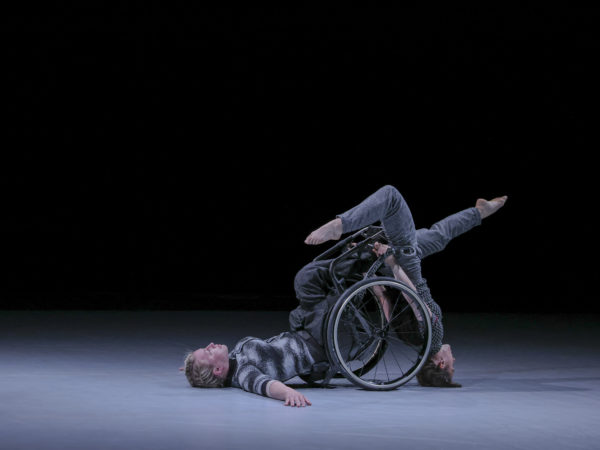 Two white female dancers. The first is a wheelchair user with her back on the ground and legs facing upwards. The other does a headstand against the wheels of her her chair with both her legs off at opposite jaunty angles.