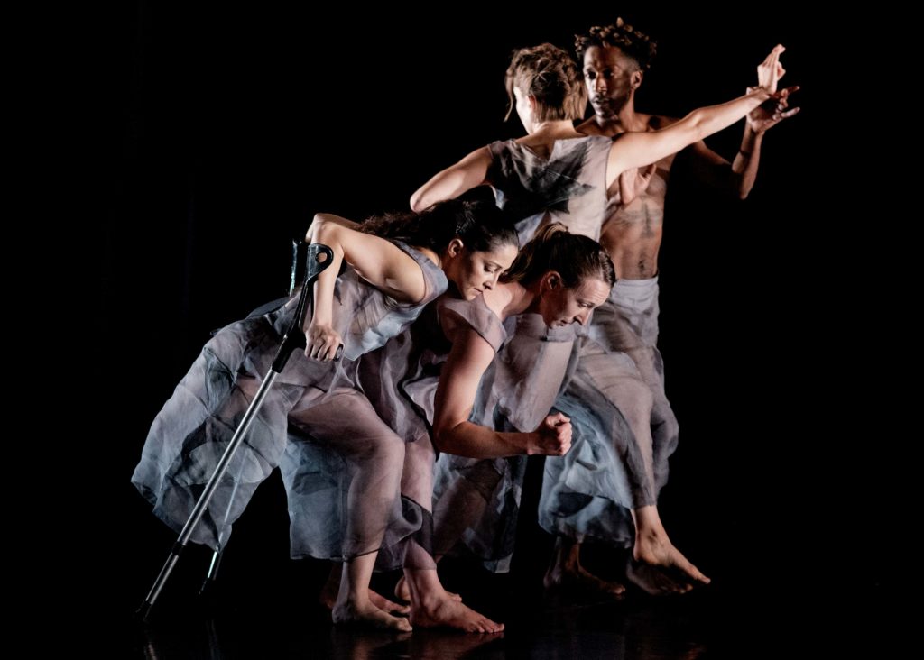 4 dancers are on an all-black stage. They wear loose-fitting sheer grey clothes. The 2 female dancers in front are side-on and crouched down, bending at the knee and upper bodies tilted forward. The front-most dancer holds 2 crutches behind her and rests her cheek on the dancer directly behind her, who holds her front arm at a right angle, as if ready to run. Behind them is another female dancer who is facing away so her back is visible. She holds her right arm straight out to her side, whilst her left arm is held out to the side and bent at the elbow. She is looking at her elbow. A male dancer faces her, and is about to hold onto her right wrist. He is looking at her face.