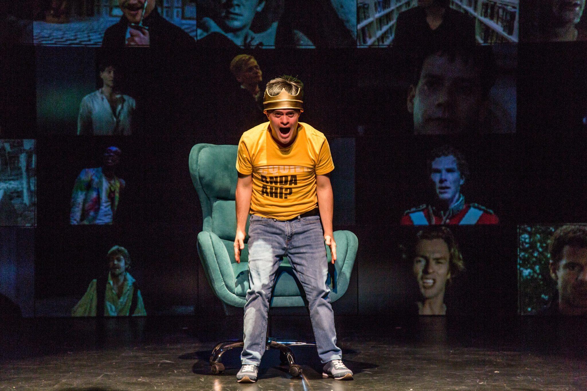 A still image from Hamlet. A performer stands on a stage in front of a teal armchair wearing a yellow t-shirt and blue jeans, with a heavy looking crown perched atop their head. They are facing the audience with their mouth open, as if caught in the middle of a passionate yell. Behind them is a large screen filled with multiple depictions of Shakespearian actors from movies and theatre.