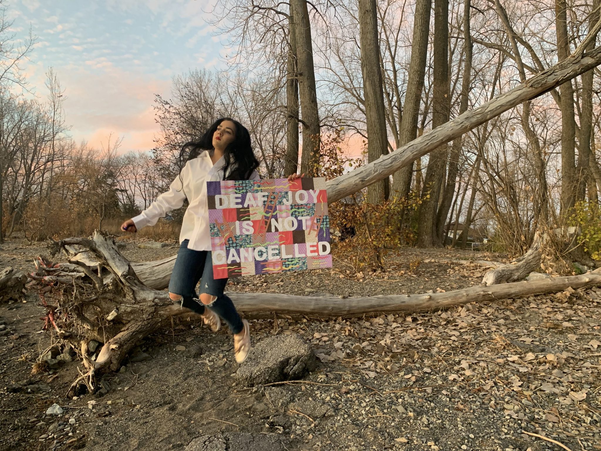 Photograph of Syra Hassan in the middle of a jump in a wooded area. In her right hand, she holds a camera remote control and in her left hand, a cardboard-sized sign decorated with a single hand that reads "Deaf joy is not cancelled"