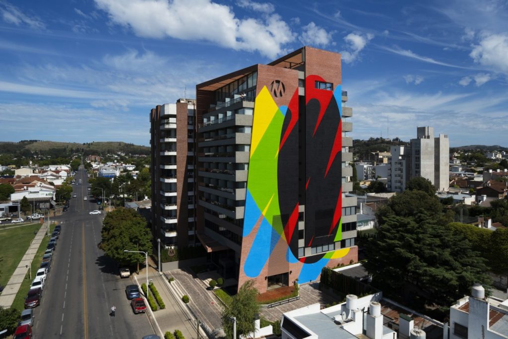 Photo of the side of a tall, brick apartment building in a mid-sized city. On the side of the building is painted a colourful mural, broad strokes of yellow, blue, green, red and black